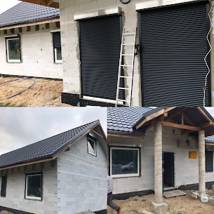 Szare rolety zewnętrzne zintegrowane z ramą okienną w nowym, surowym budynku z białego bloczka. Widoczna drabina oparta o ścianę przy jednej z rolet. Budynek w stanie surowym otwartym, bez elewacji.