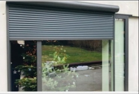 Zewnętrzna roleta zwijana w kolorze antracytowym, częściowo opuszczona na dużym oknie przesuwnym, z widocznym odbiciem otoczenia i choinki w szybie.