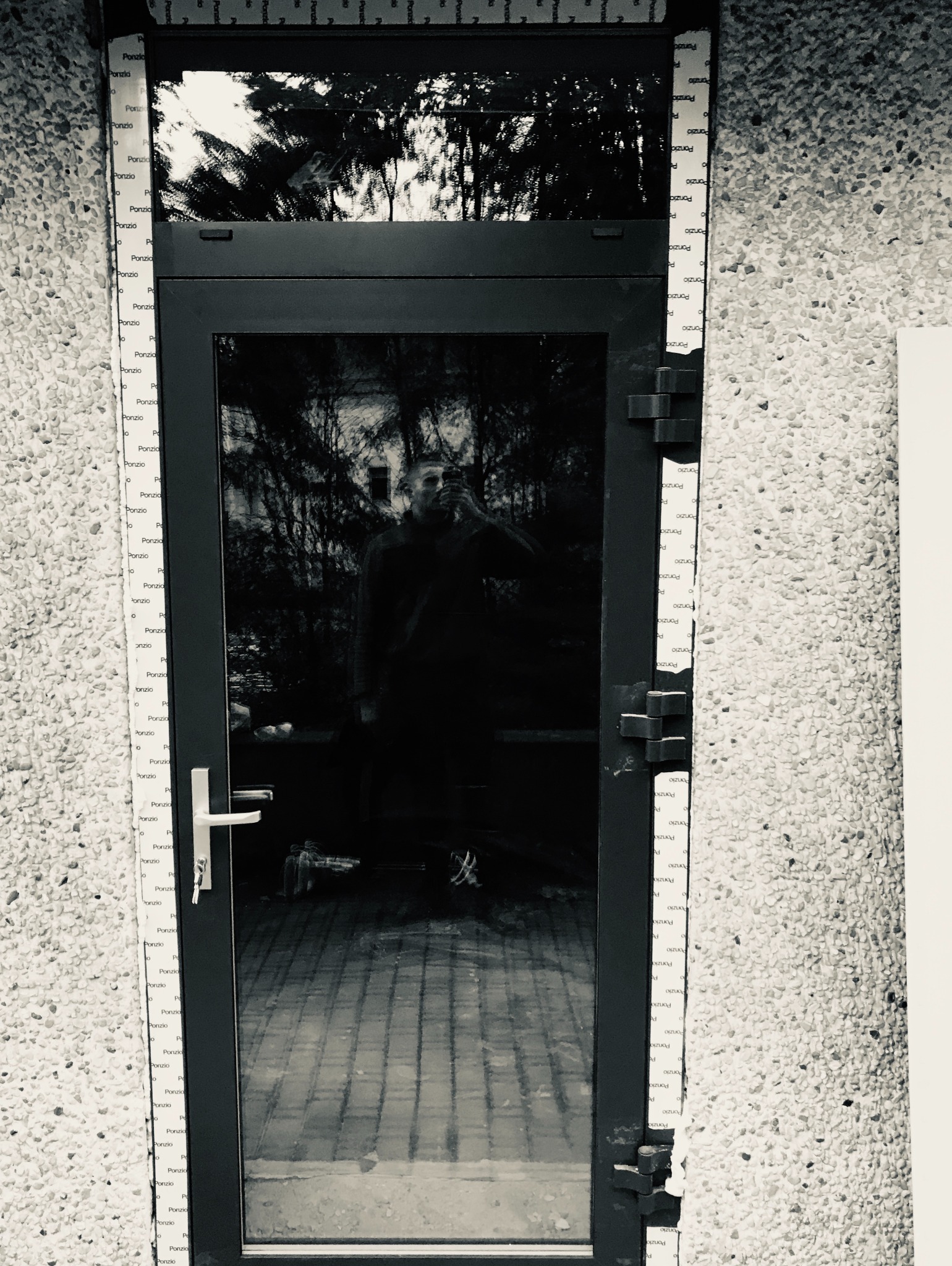 Czarne drzwi wejściowe z widocznym odbiciem osoby robiącej zdjęcie, otoczone ścianą z jasnego kamienia, zabezpieczone folią ochronną z logo producenta.