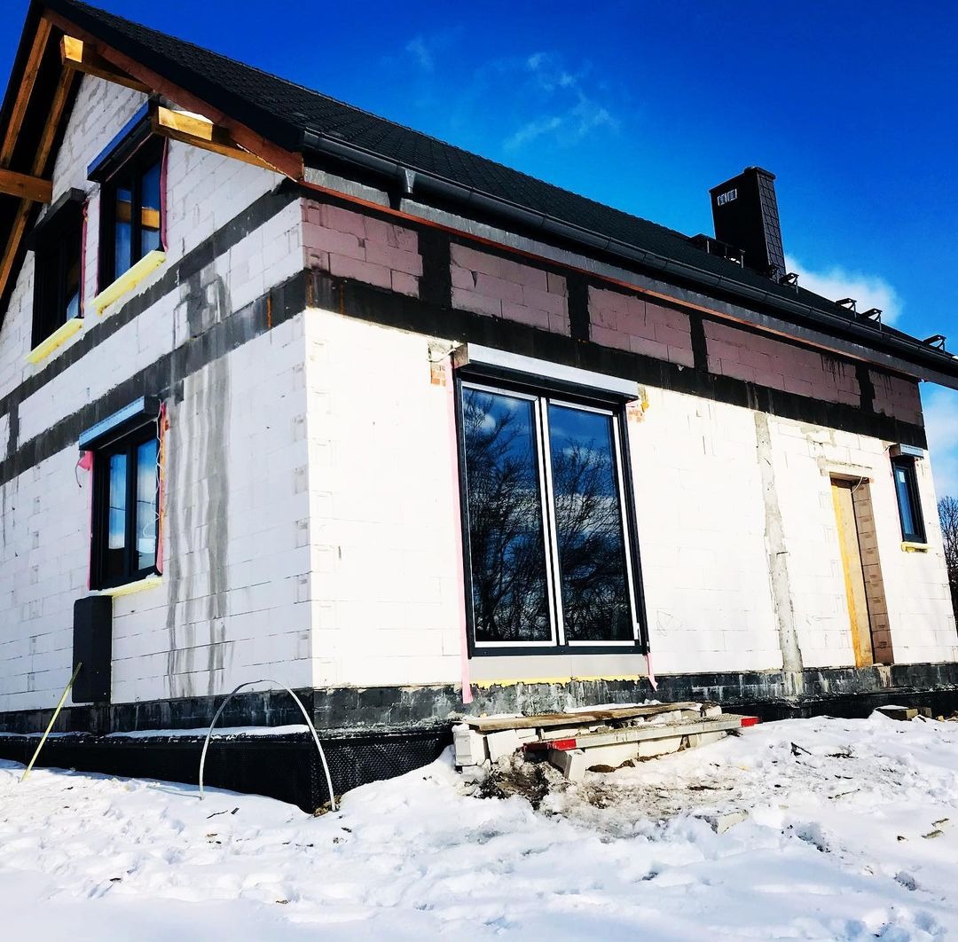 Dom w trakcie budowy w zimowym krajobrazie, z zamontowanymi oknami PCV z roletami zewnętrznymi i przygotowanymi otworami na kolejne okna.