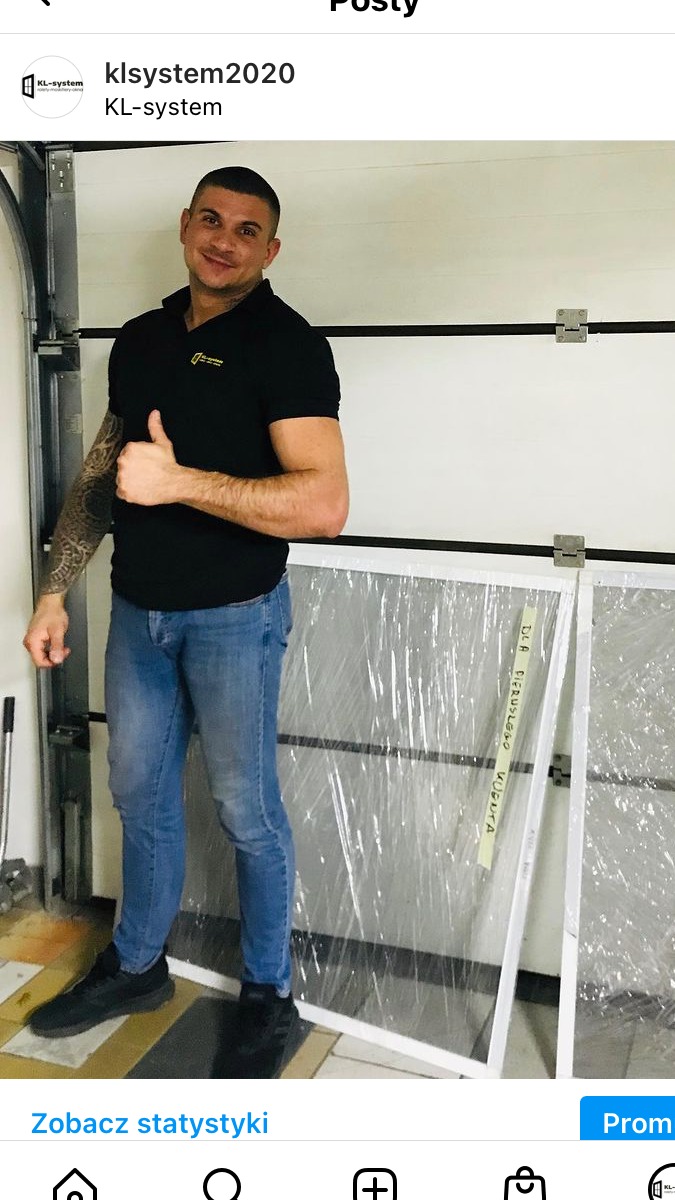 Mężczyzna w czarnej koszulce polo z logo firmy, jeansach i czarnych butach, stoi na tle garażu z dwoma zapakowanymi moskitierami i uniesionym kciukiem.