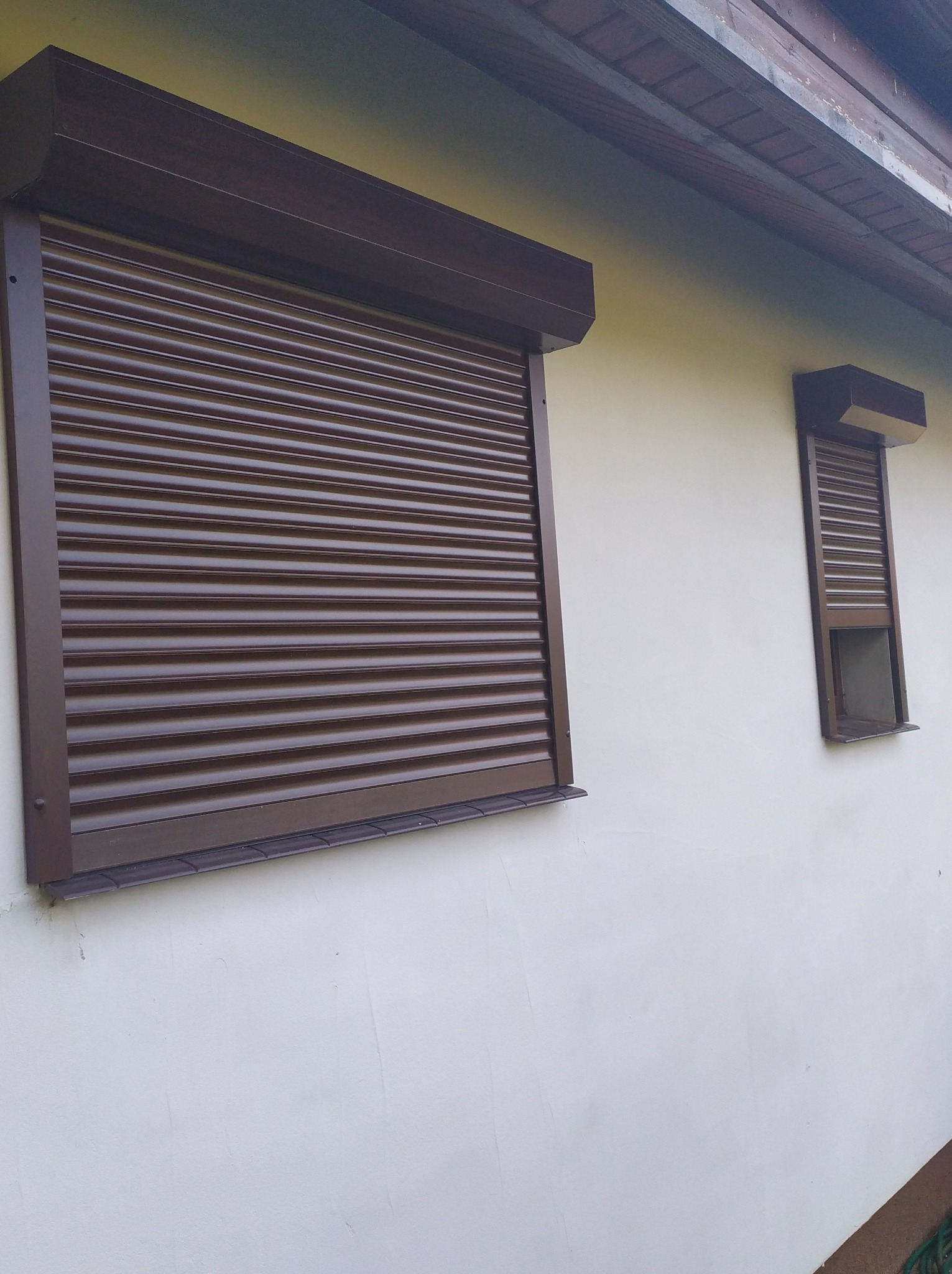 Brązowe rolety zewnętrzne zamontowane na dwóch oknach w jasnym budynku, jedno okno całkowicie zasłonięte, drugie częściowo otwarte.