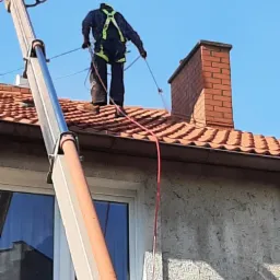 Pracownik w uprzęży, z podnośnika koszowego, myje czerwoną dachówkę spadzistego dachu domu jednorodzinnego, widoczny komin z cegły i fragment elewacji.