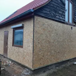 Dom w trakcie przygotowania do ocieplenia elewacji, widoczne płyty OSB na ścianach, fragment starej drewnianej elewacji i zwinięty kabel na ziemi.