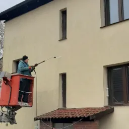 Pracownik w podnośniku koszowym czyści elewację budynku za pomocą myjki ciśnieniowej. Widoczny fragment elewacji przed i po czyszczeniu, ukazujący różnicę w kolorze.