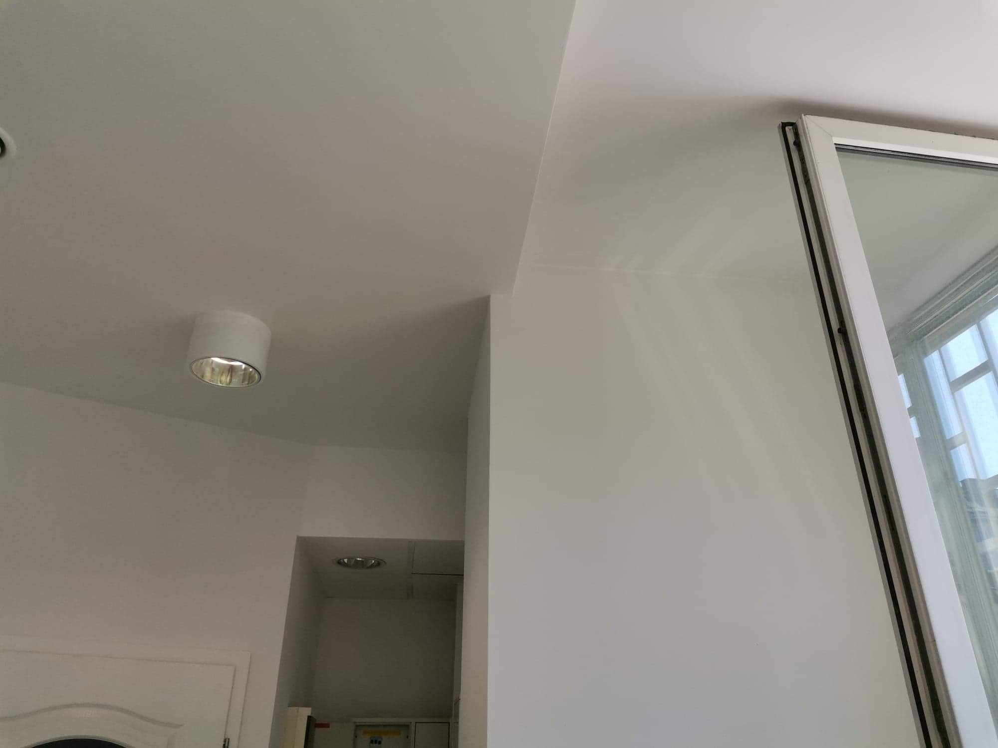 Wnętrze po malowaniu na biało, widoczny fragment sufitu z okrągłą lampą, biała ściana z oknem w białej ramie i fragment drzwi w tle.