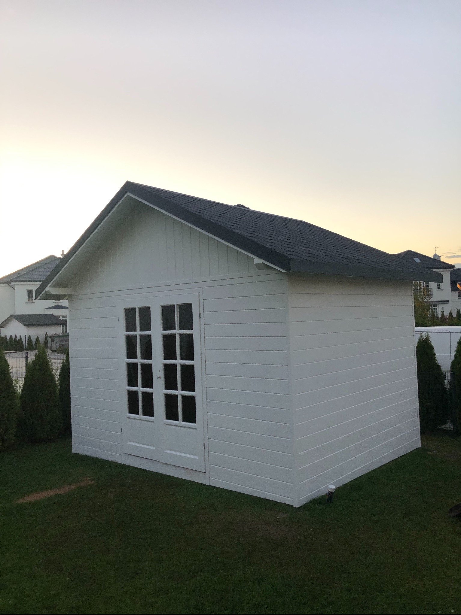 Biały, drewniany domek narzędziowy z dwuskrzydłowymi przeszklonymi drzwiami, na tle trawnika i zabudowań mieszkalnych o zmierzchu.