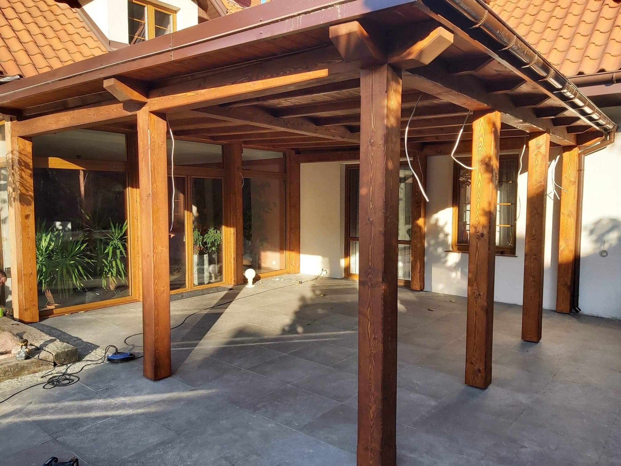 Drewniana konstrukcja zadaszenia tarasu przylegająca do domu z widocznymi oknami i roślinami doniczkowymi wewnątrz, na podłodze leżą narzędzia.