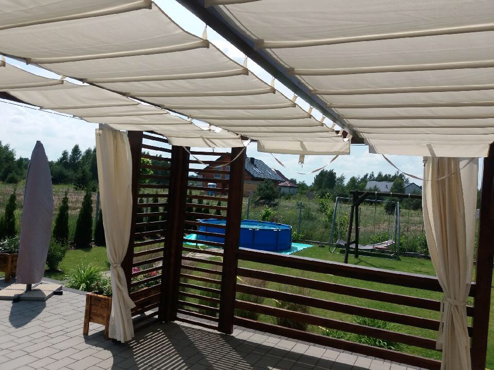 Drewniana pergola z beżowym, płóciennym zadaszeniem i zasłonami, widok na ogród z basenem i huśtawką.
