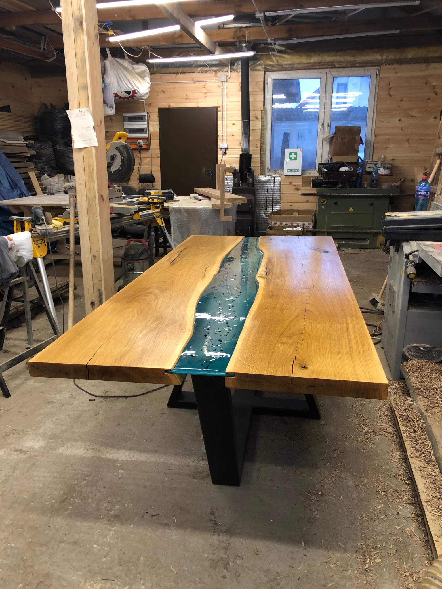 Stół z blatem z dwóch desek połączonych żywicą epoksydową imitującą rzekę, na metalowych nogach, w warsztacie stolarskim.