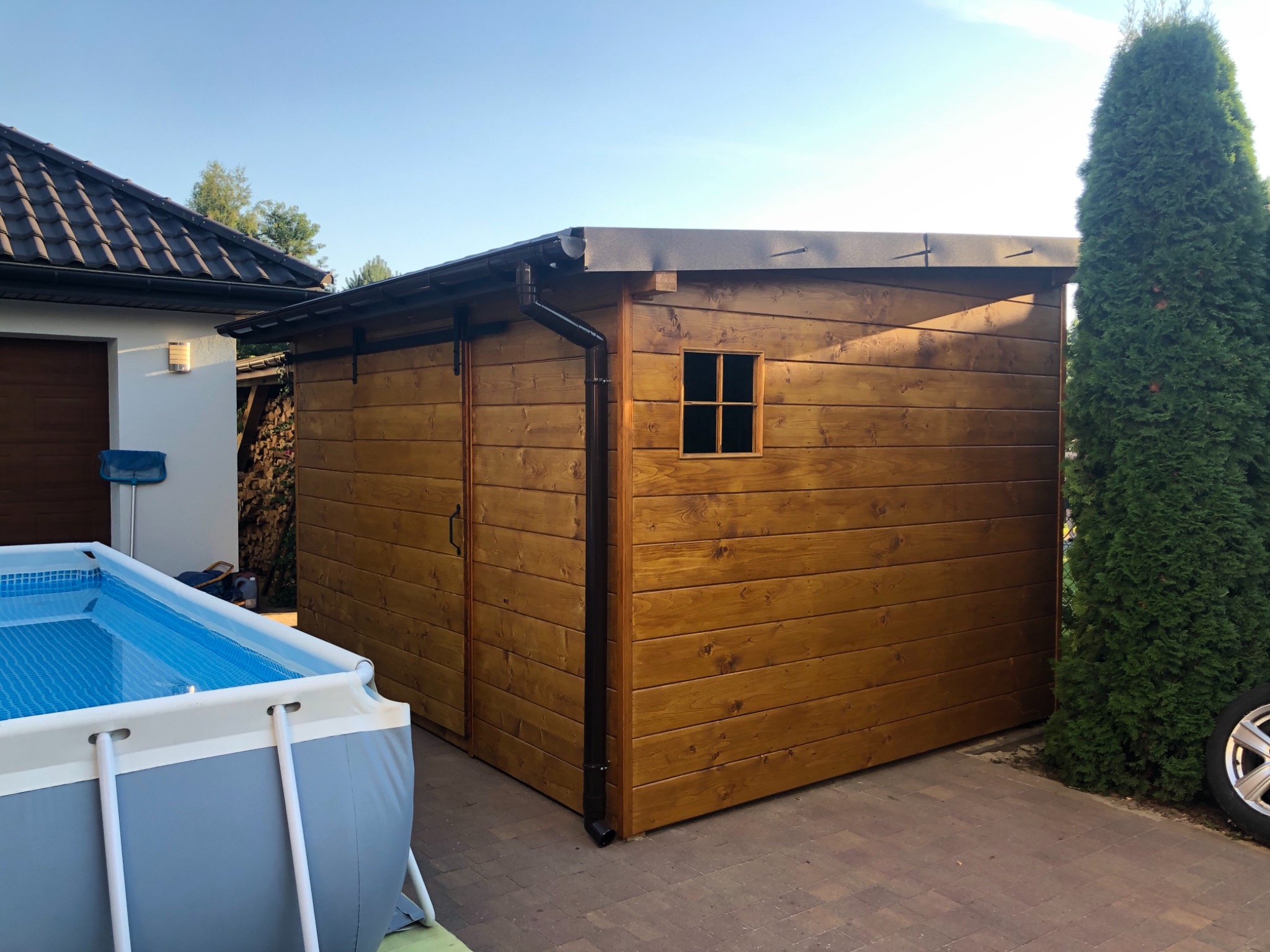 Drewniany domek narzędziowy z oknem i rynną, stojący obok basenu i tuj, na tle domu z brązowym dachem.