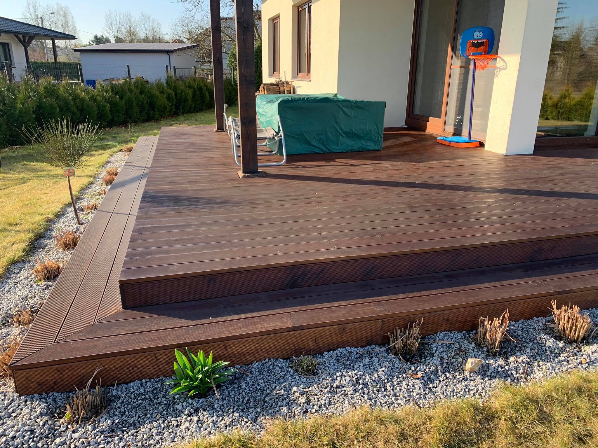 Nowoczesny taras z ciemnego drewna z dwoma stopniami, obłożony szarym żwirem i młodymi roślinami, widok na dom z plastikowym koszem do koszykówki