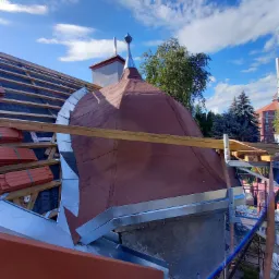 Kopuła dachu pokryta papą i obłożona blachą, widoczne elementy więźby dachowej, rynny i rusztowanie na tle błękitnego nieba.