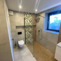 Nowoczesna łazienka z kabiną prysznicową typu walk-in, toaleta podwieszana, jasne płytki imitujące beton, drewnopodobna podłoga w strefie prysznica i akcent roślinny na ścianie.
