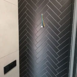 Ciemnoszara ściana w łazience wyłożona płytkami ułożonymi w jodełkę, z wystającymi przewodami i czarnym panelem sterującym spłuczką.