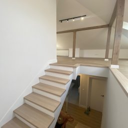 Margo sp.z.o.o - Jasne drewniane schody prowadzące na antresolę z drewnianymi belkami, widoczne świeżo ułożone panele podłogowe i pomalowane na biało ściany.