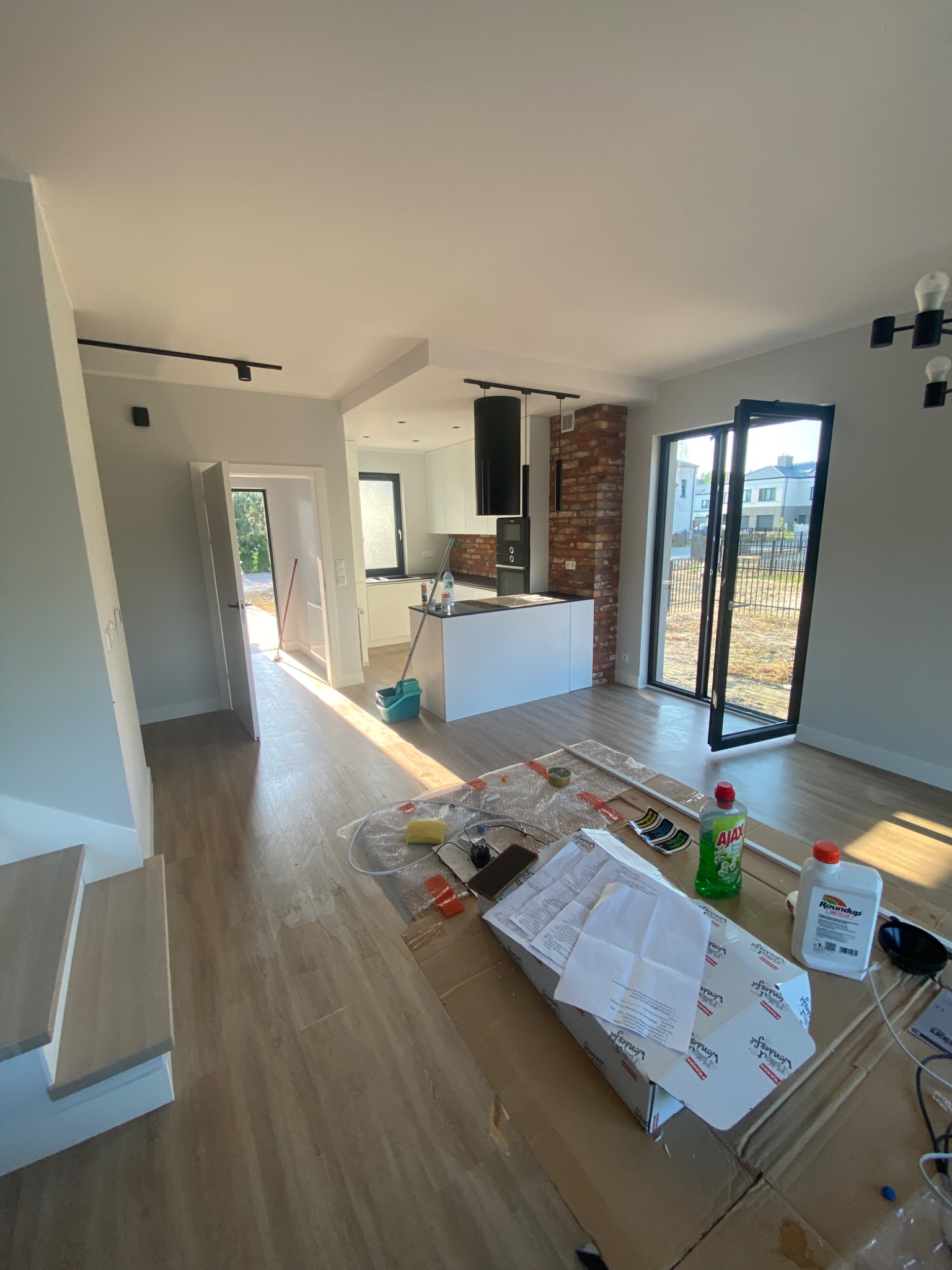 Wnętrze w trakcie prac wykończeniowych: widok na salon z aneksem kuchennym, podłoga częściowo zabezpieczona folią, widoczne środki czystości i opakowania po materiałach budowlanych, ceglana ściana...