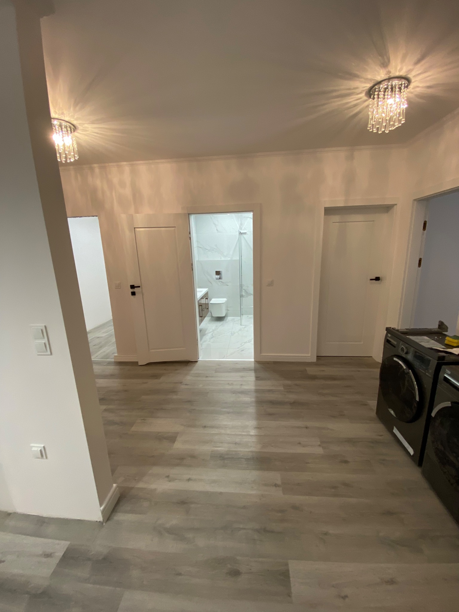 Jasne, przestronne wnętrze mieszkania po remoncie, z panelami podłogowymi imitującymi drewno, widok na łazienkę z marmurowymi płytkami i toaletą, oraz nowoczesne lampy sufitowe.