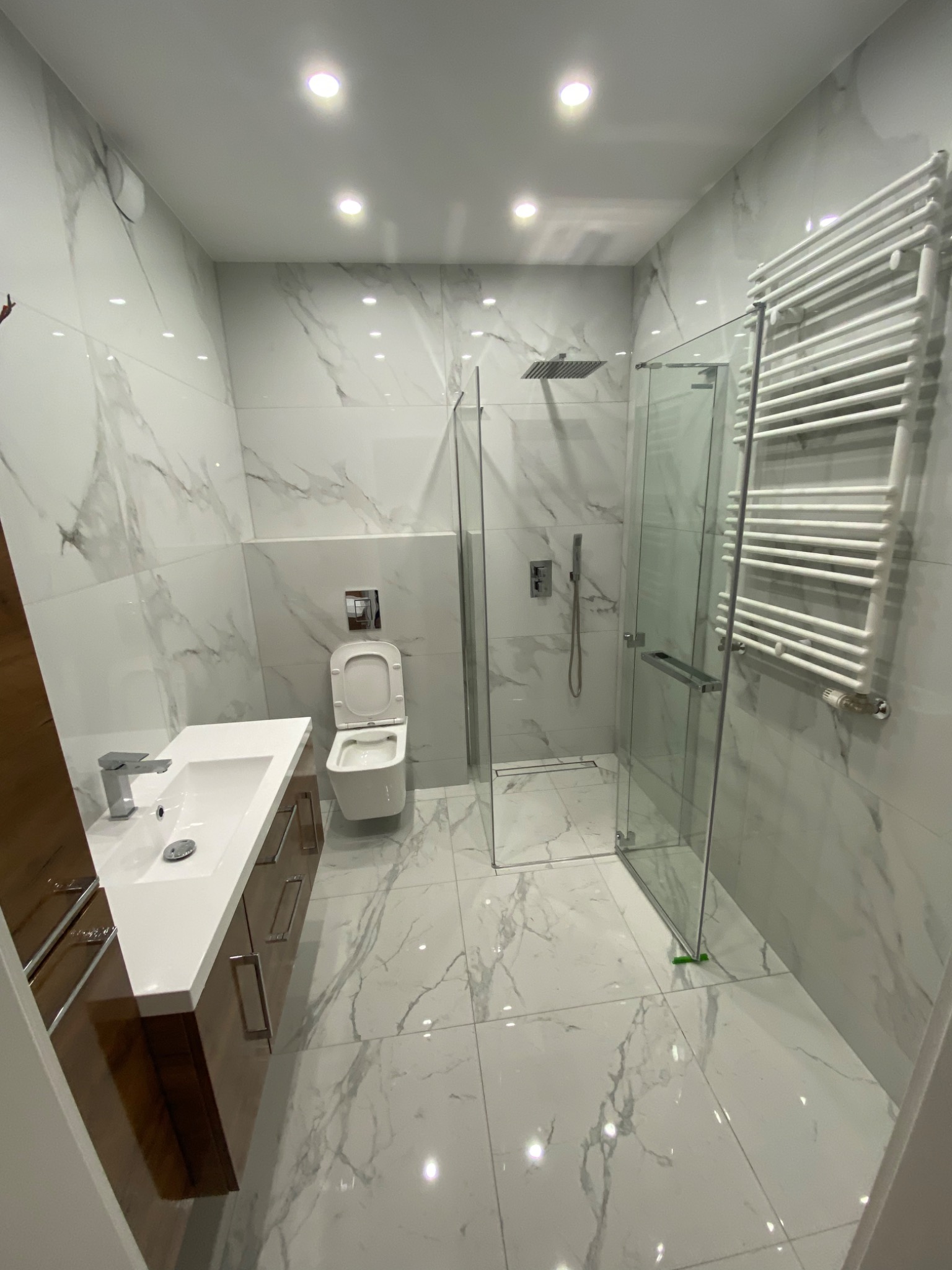 Nowoczesna łazienka z marmurowymi płytkami, kabiną prysznicową typu walk-in, podwieszaną toaletą i umywalką z szafką w kolorze drewna.