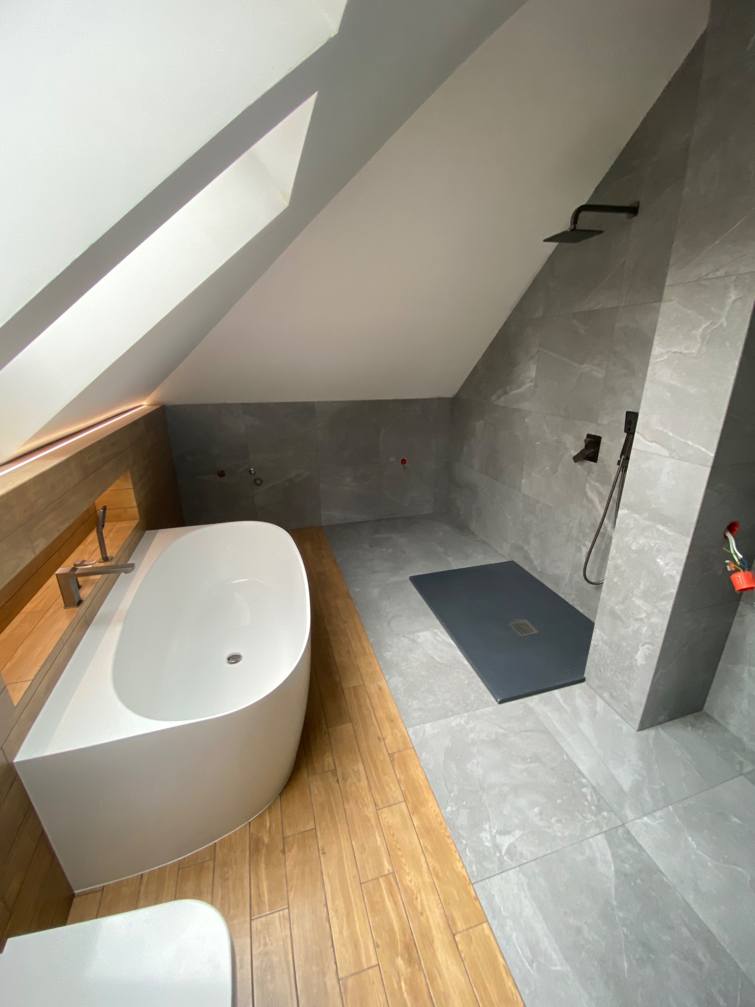 Nowoczesna łazienka na poddaszu z białą wanną, drewnianą zabudową i szarą strefą prysznicową z czarną armaturą, wykończona płytkami imitującymi marmur.