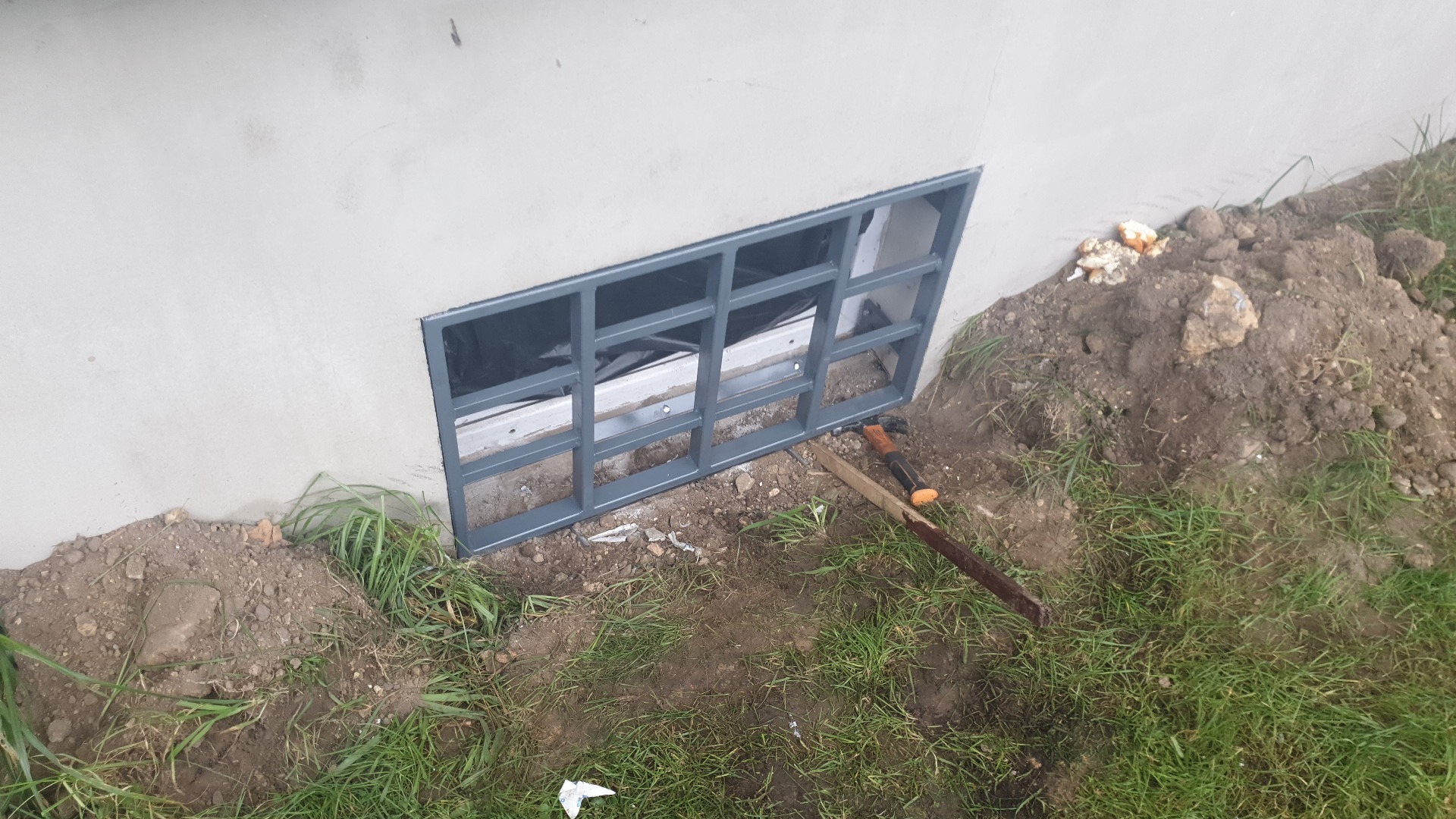 Szara metalowa krata okienna osadzona w fundamencie budynku w Żorach, widoczne narzędzia i ziemia.