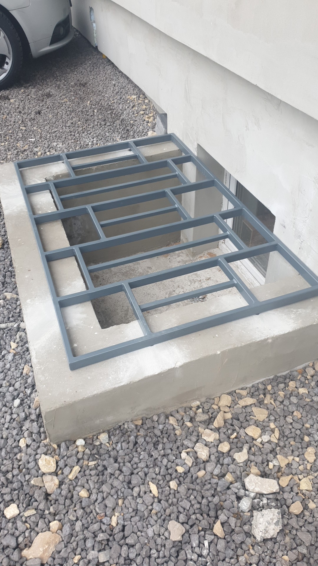 Szara, metalowa krata zabezpieczająca okno piwniczne, zamontowana w betonowej wnęce przy ścianie budynku, otoczona żwirem.