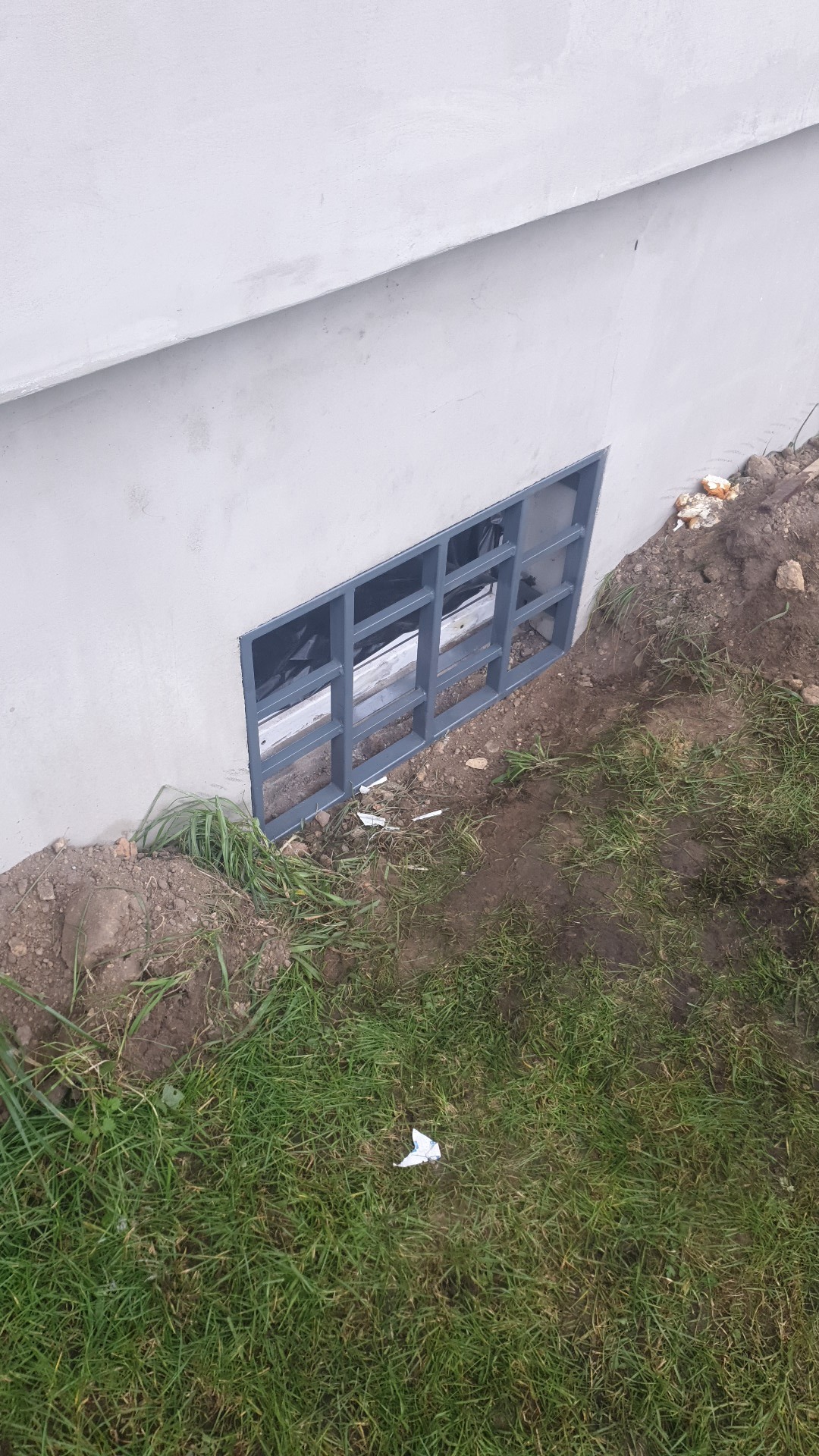 Metalowa krata okienna zabezpieczająca piwnicę wkomponowana w szarą ścianę budynku, widoczna trawa i ziemia.