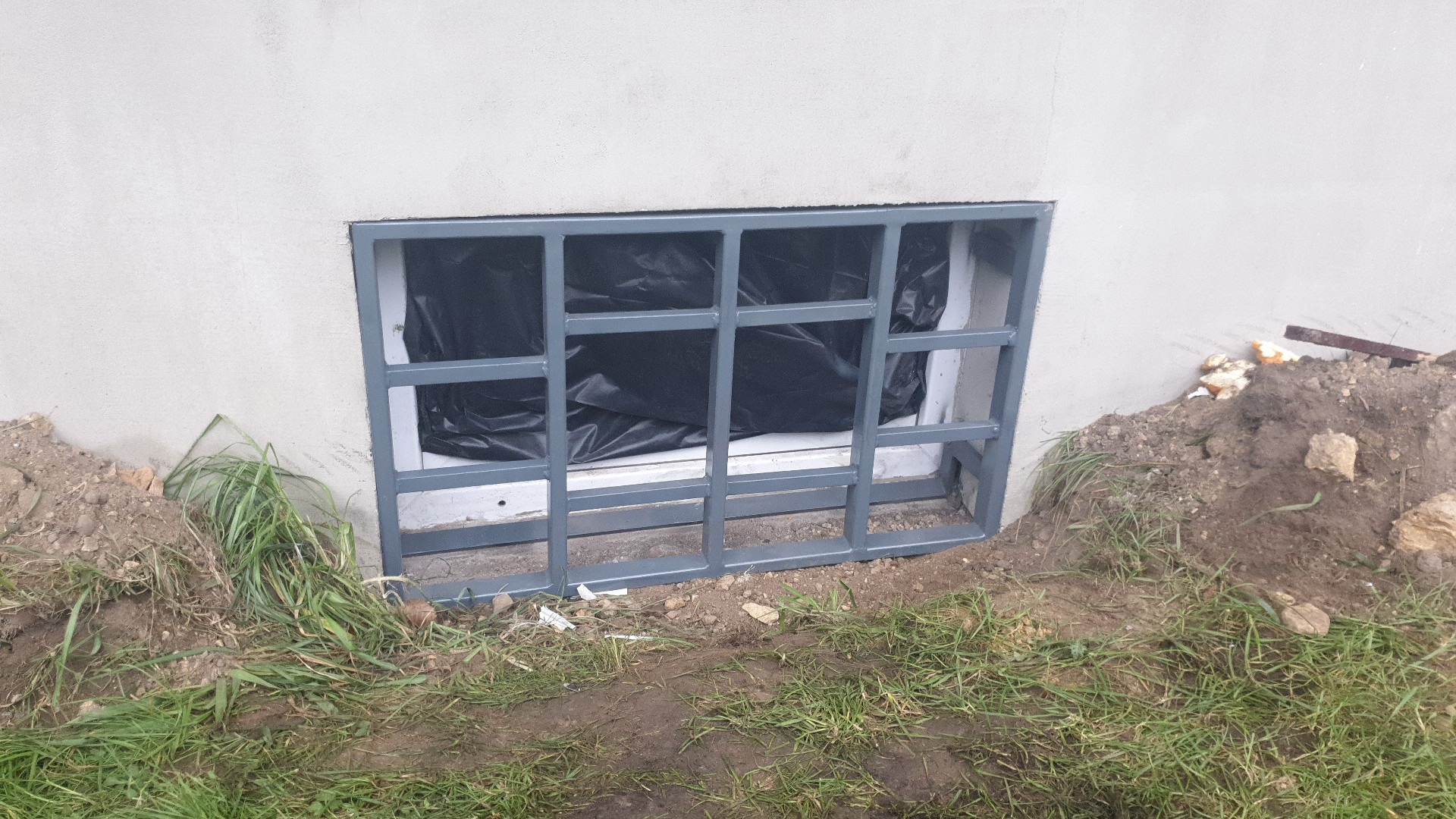 Metalowa krata okienna zabezpieczająca piwniczne okno, pomalowana na szaro, z czarną folią w tle, otoczona ziemią i trawą.