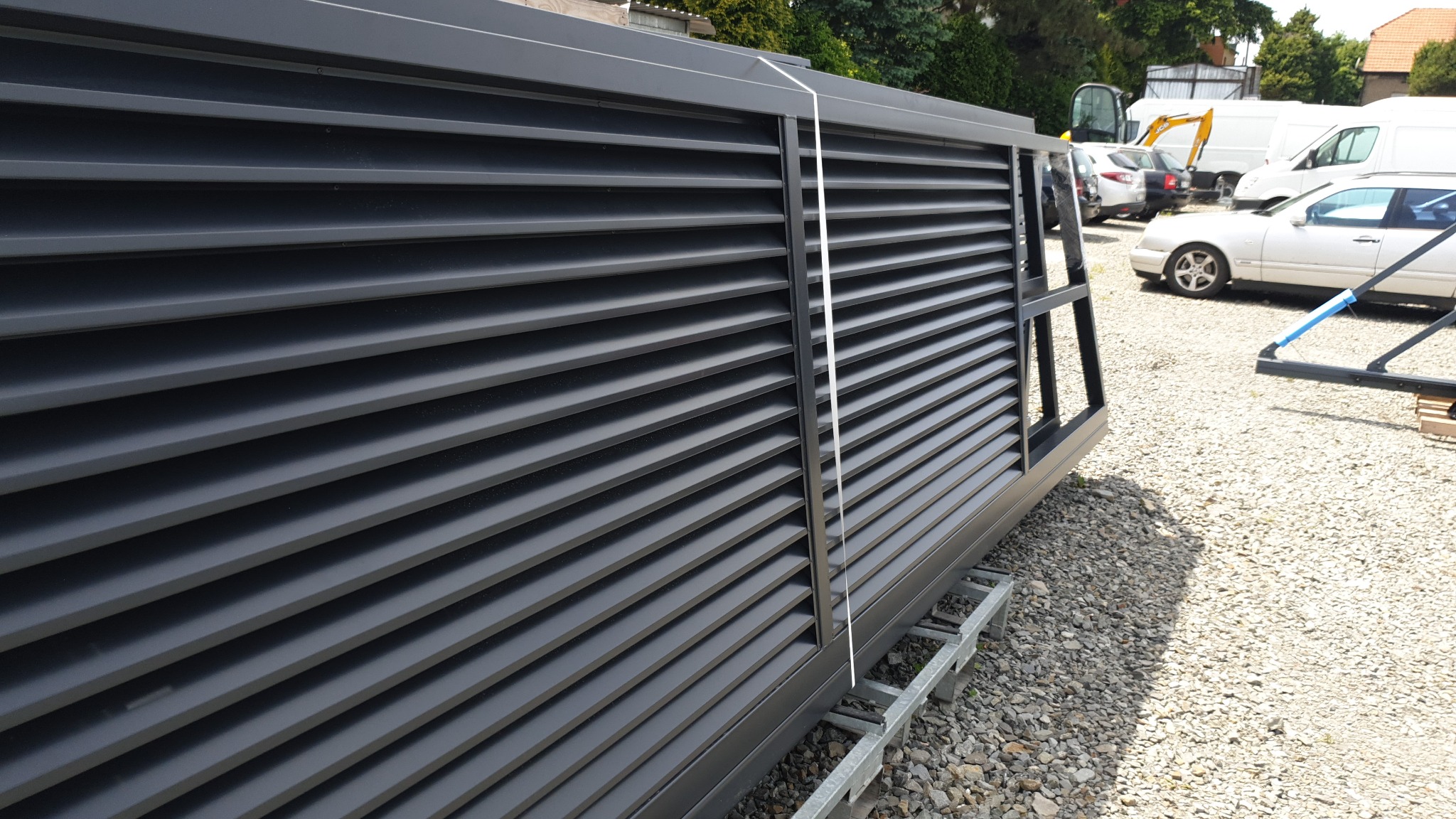 Nowoczesna brama przesuwna z poziomych, czarnych lameli, jeszcze nie zamontowana, leżąca na metalowych profilach na żwirowym placu.