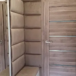 Kącik do siedzenia z tapicerowanymi panelami ściennymi w kolorze beżowym, z siedziskiem i szafką wnękową, obok drewnianych drzwi z metalową klamką, na marmurowej podłodze.