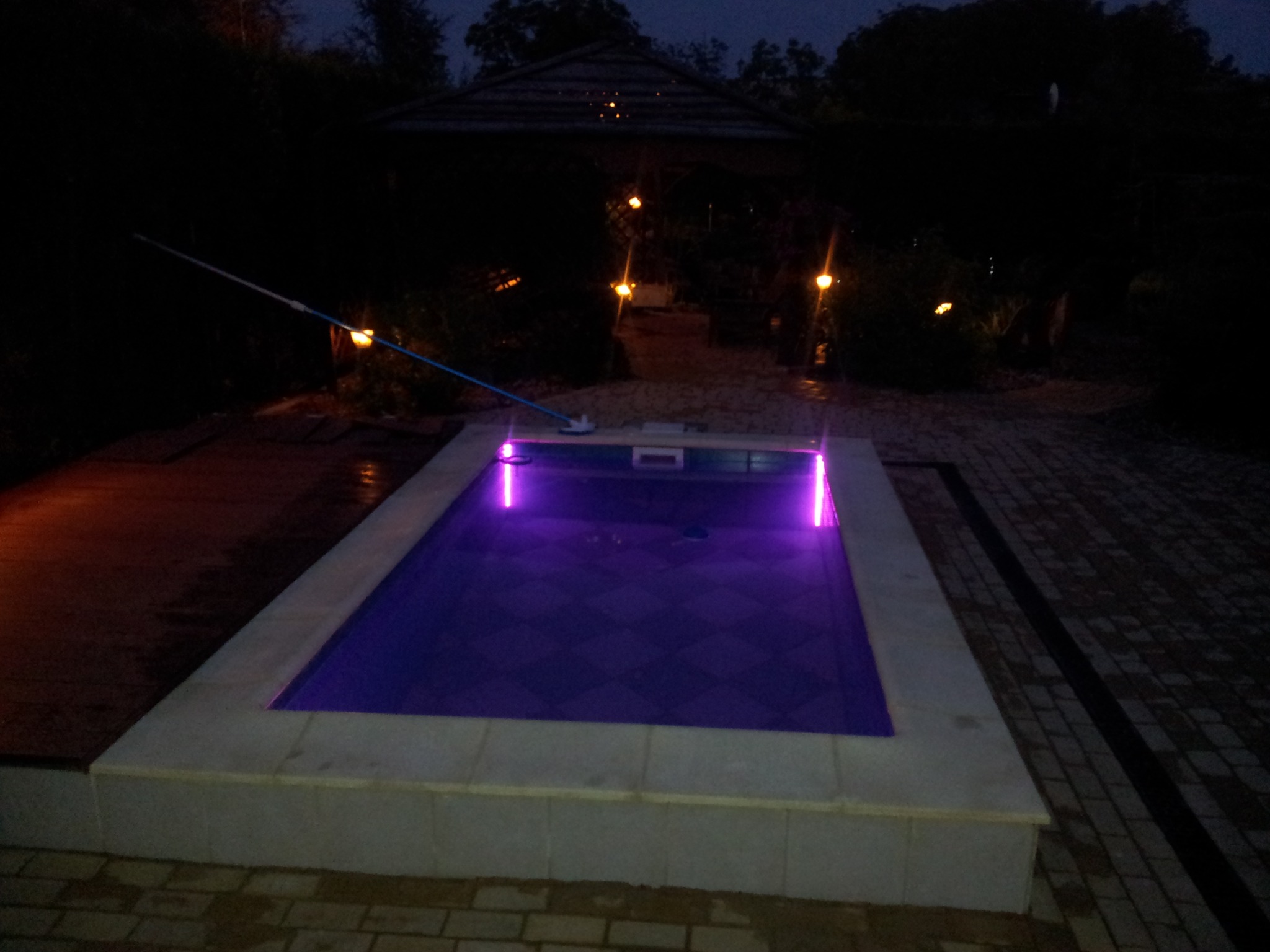 Oświetlony basen z purpurowym światłem w ogrodzie nocą, widoczny kij do czyszczenia i altana z lampkami.