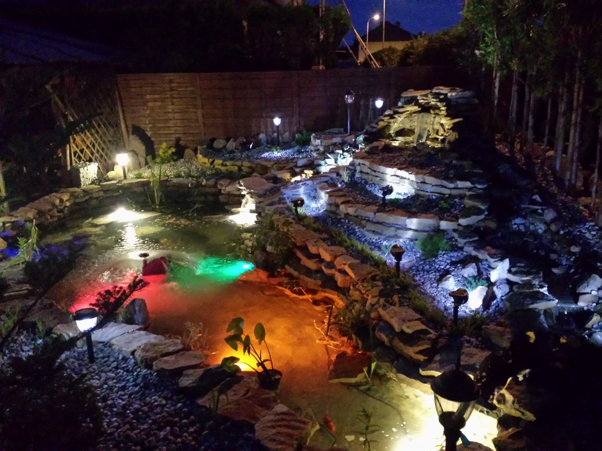 Oświetlony nocą, wielopoziomowy staw ogrodowy z kaskadami, obłożony kamieniami i żwirem, z podwodnymi lampami w kolorach czerwonym, zielonym i pomarańczowym oraz lampami ogrodowymi wzdłuż ścieżki.
