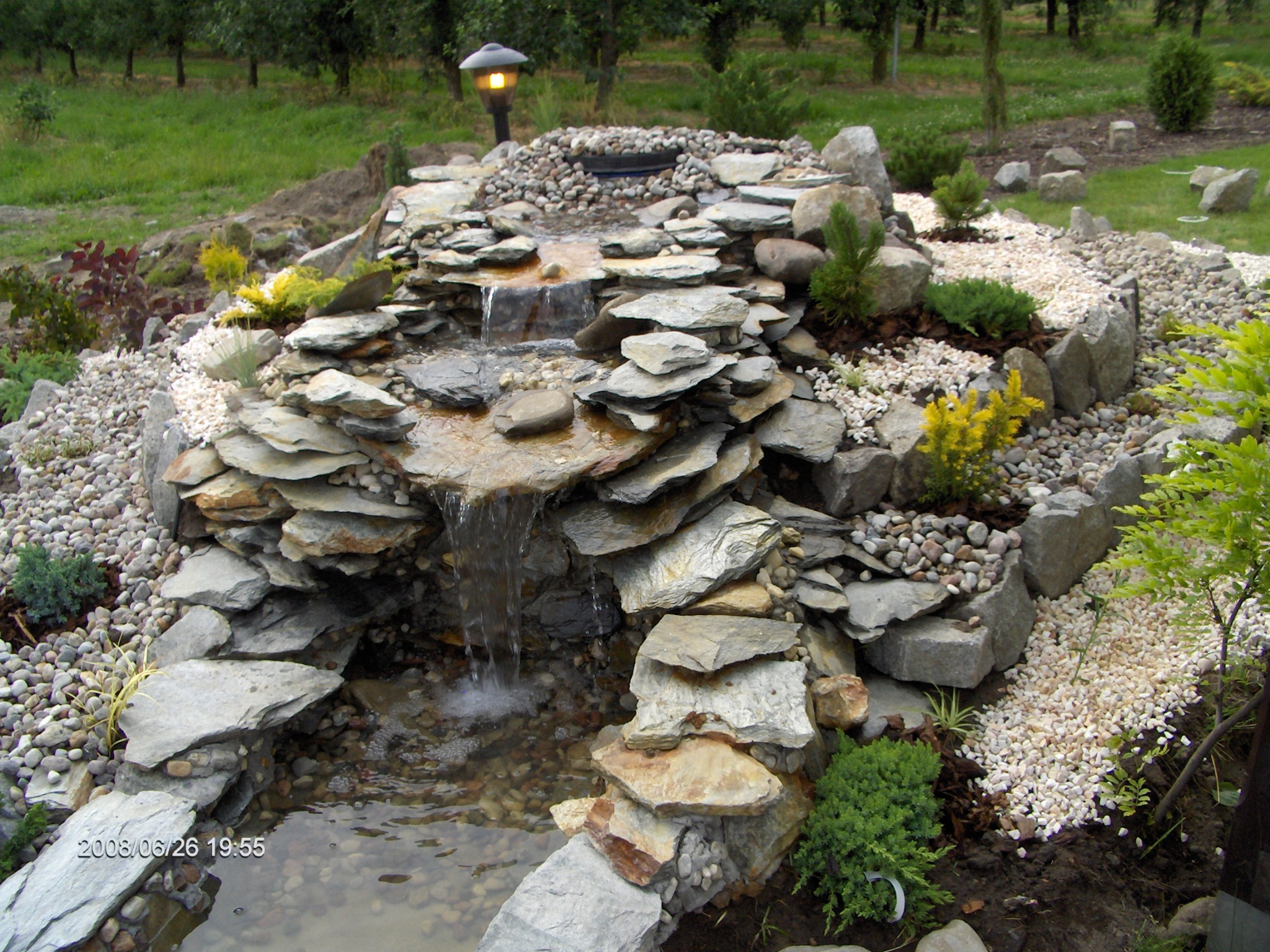 Ogród skalny z kaskadą wodną, oświetlony lampą ogrodową, otoczony zielenią i białym żwirem.