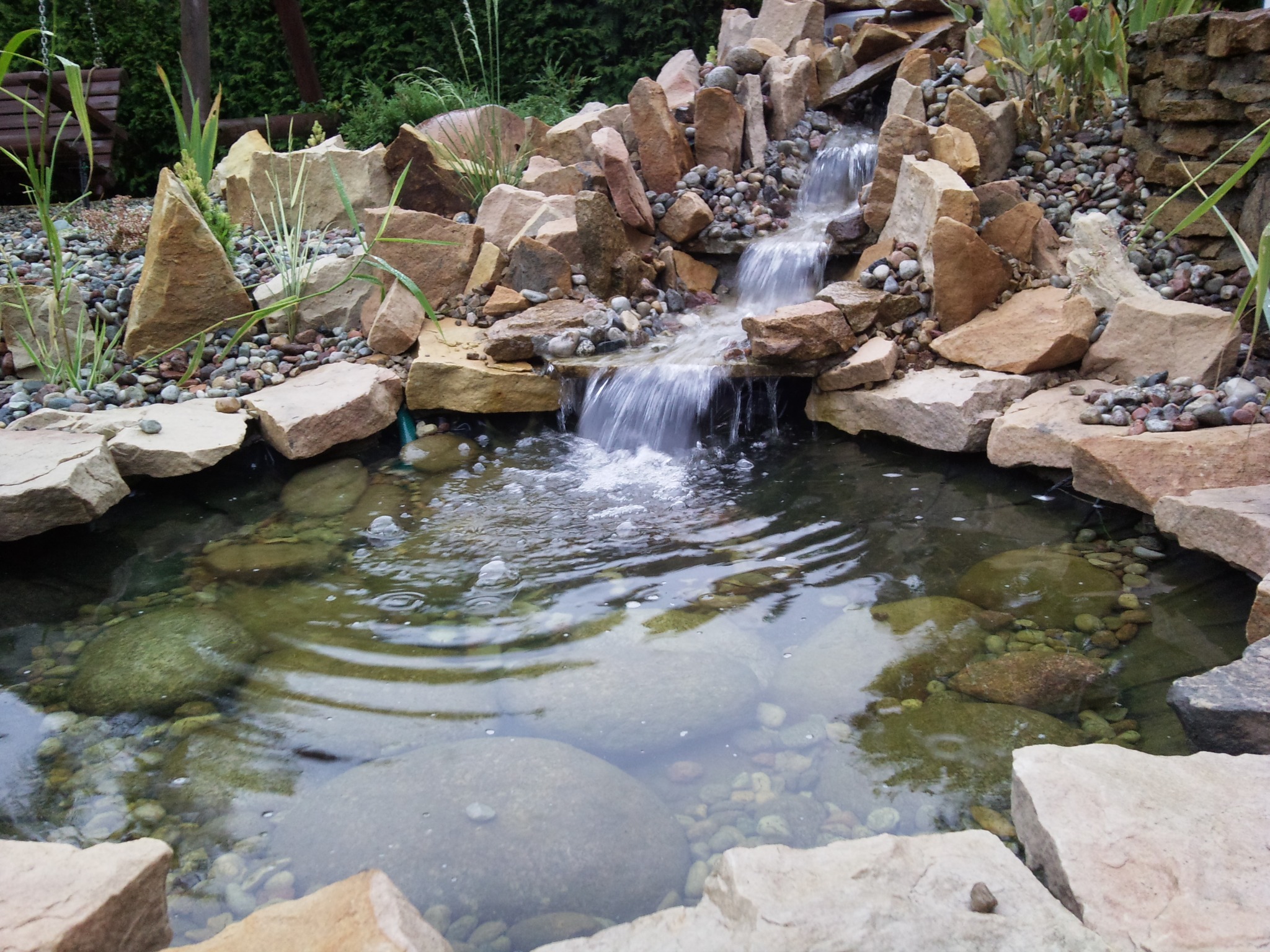 Kaskada wodna zbudowana z kamieni o różnej wielkości, tworząca mały staw z widocznymi kamieniami na dnie i roślinnością wokół.