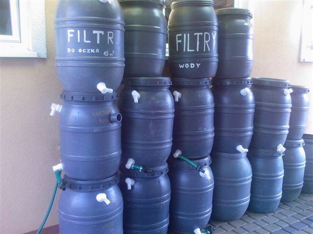 Instalacja filtrów do wody w zbiornikach z tworzywa sztucznego, niektóre z napisami 'FILTR do oczka 45m3' i 'FILTRY wody', z kranikami i wężami, ustawionych jeden na drugim.