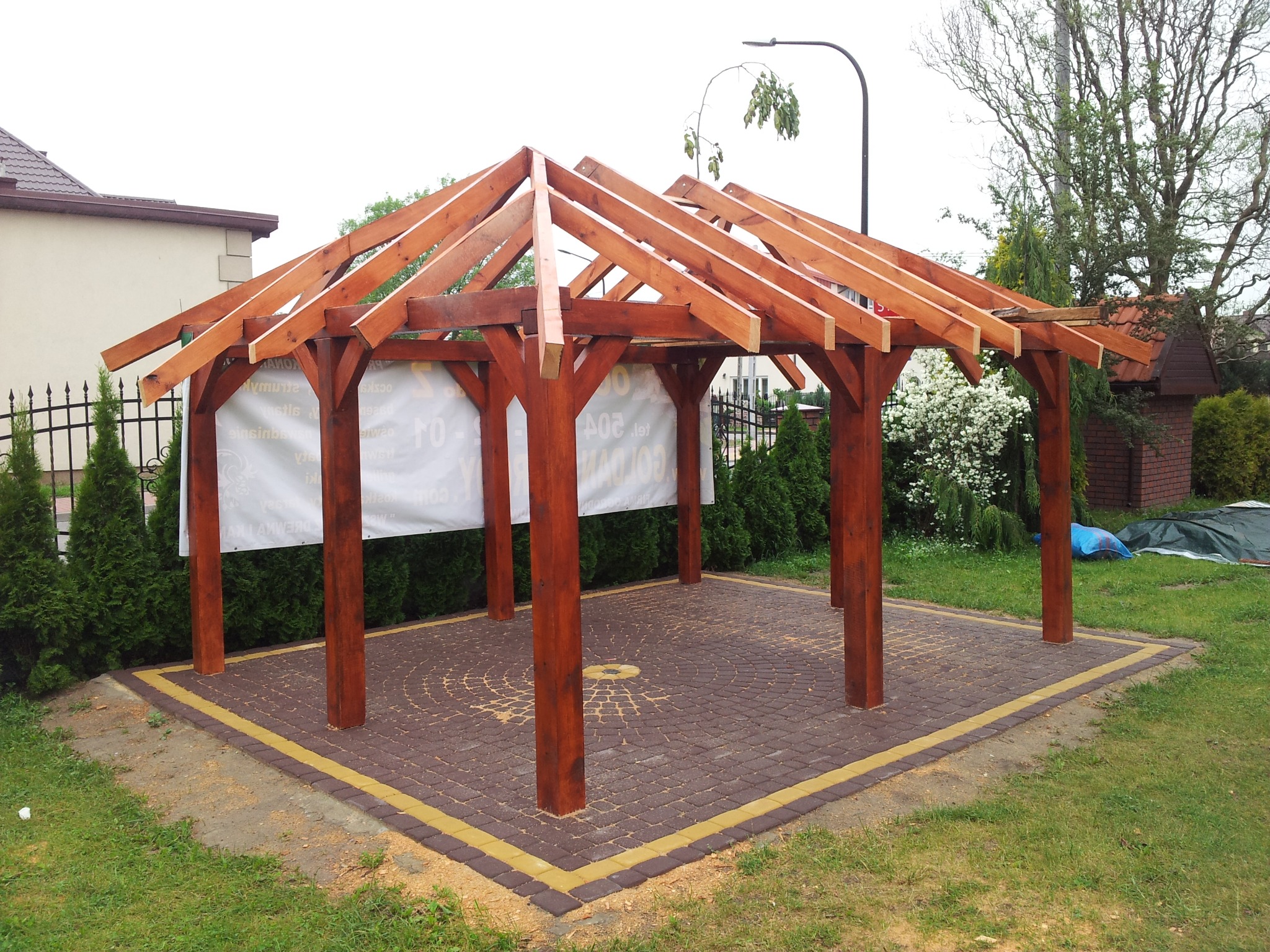 Drewniana konstrukcja altany ogrodowej w trakcie budowy, widoczne słupy i więźba dachowa, położona na kostce brukowej, w tle zieleń ogrodu i budynek.