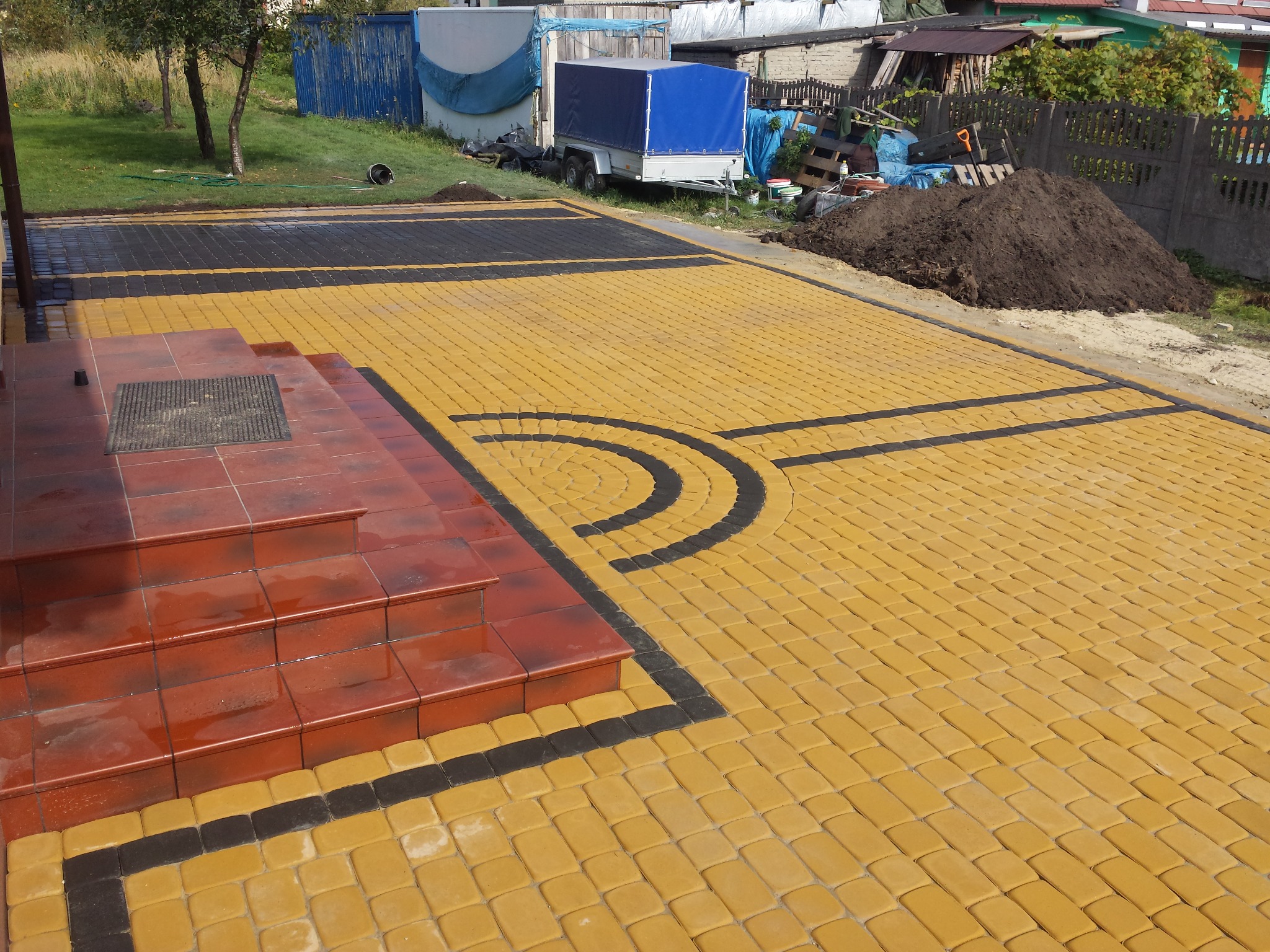 Ułożona kostka brukowa w kolorze żółtym z czarnymi elementami dekoracyjnymi przed wejściem do domu z czerwonymi schodami, w tle widoczny trawnik i przyczepa.