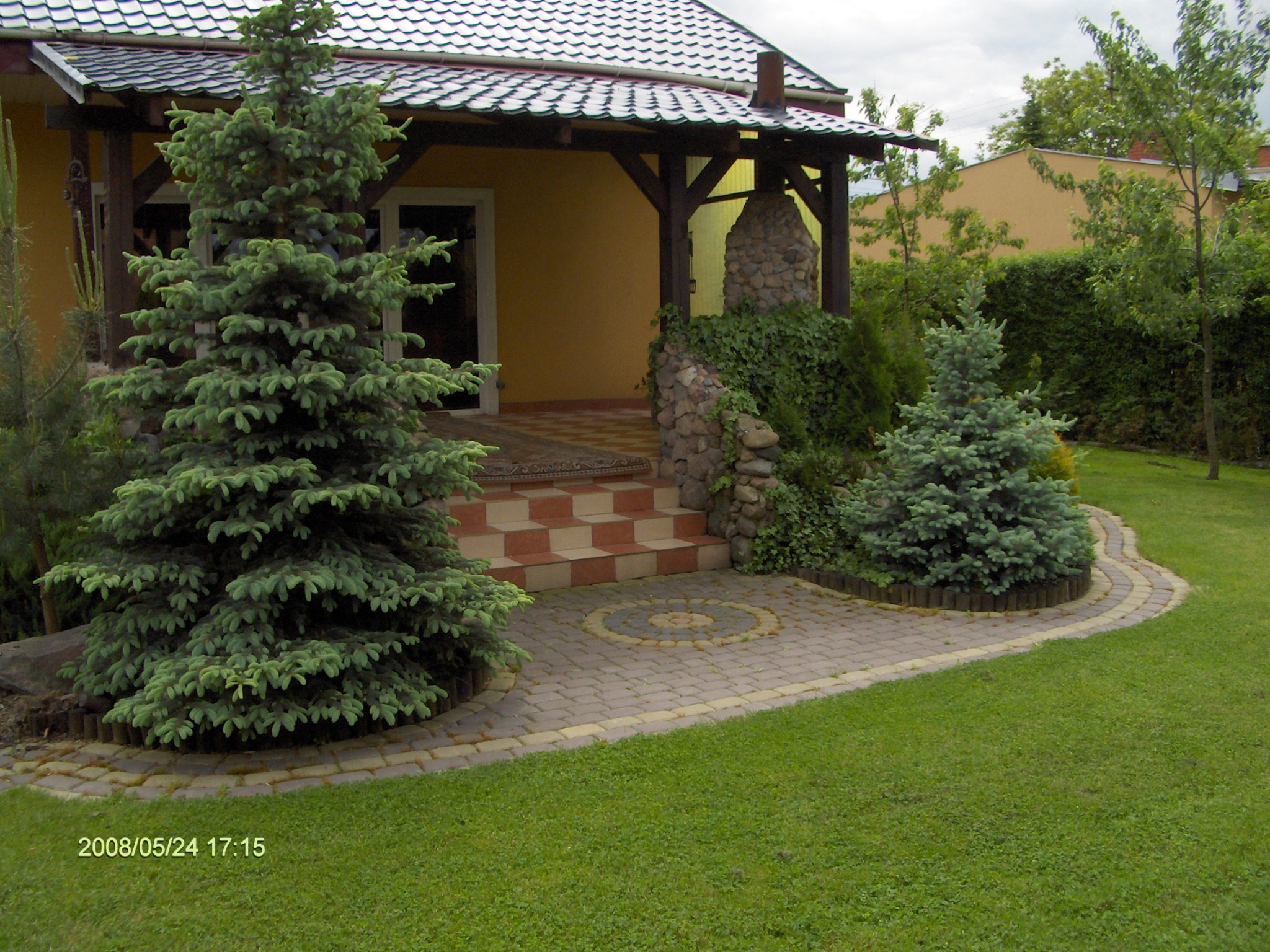 Ujęcie ogrodu z dwoma świerkami karłowymi, brukowanym chodnikiem z dekoracyjnym wzorem i schodami prowadzącymi do domu z kamienną kolumną porośniętą bluszczem.