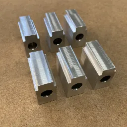 Sześć precyzyjnie obrobionych, metalowych bloków z otworem i rowkiem, ułożonych na drewnianej powierzchni, widoczne ślady obróbki.