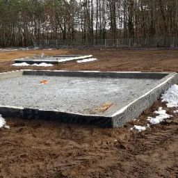 Zalany betonowy fundament z izolacją pionową i rurą w ziemi, otoczony błotnistym terenem i resztkami śniegu, w tle drzewa i ogrodzenie.