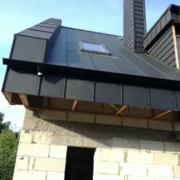 Surowy stan budynku z szarego betonu komórkowego z zamontowanym, grafitowym dachem z blachy na rąbek stojący, z oknem dachowym i kominem, widoczne drewniane elementy konstrukcji dachu.