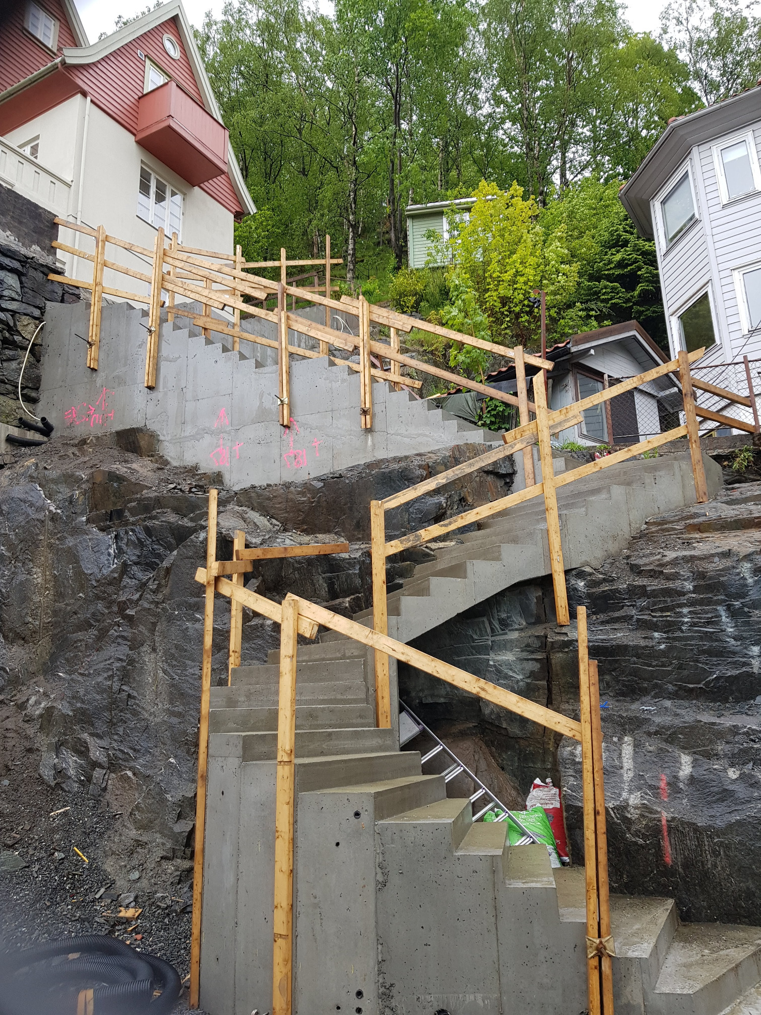 Betonowe schody zewnętrzne w trakcie budowy, prowadzące po skalistym zboczu, zabezpieczone drewnianą balustradą, z widocznymi śladami pomiarów w kolorze różowym.