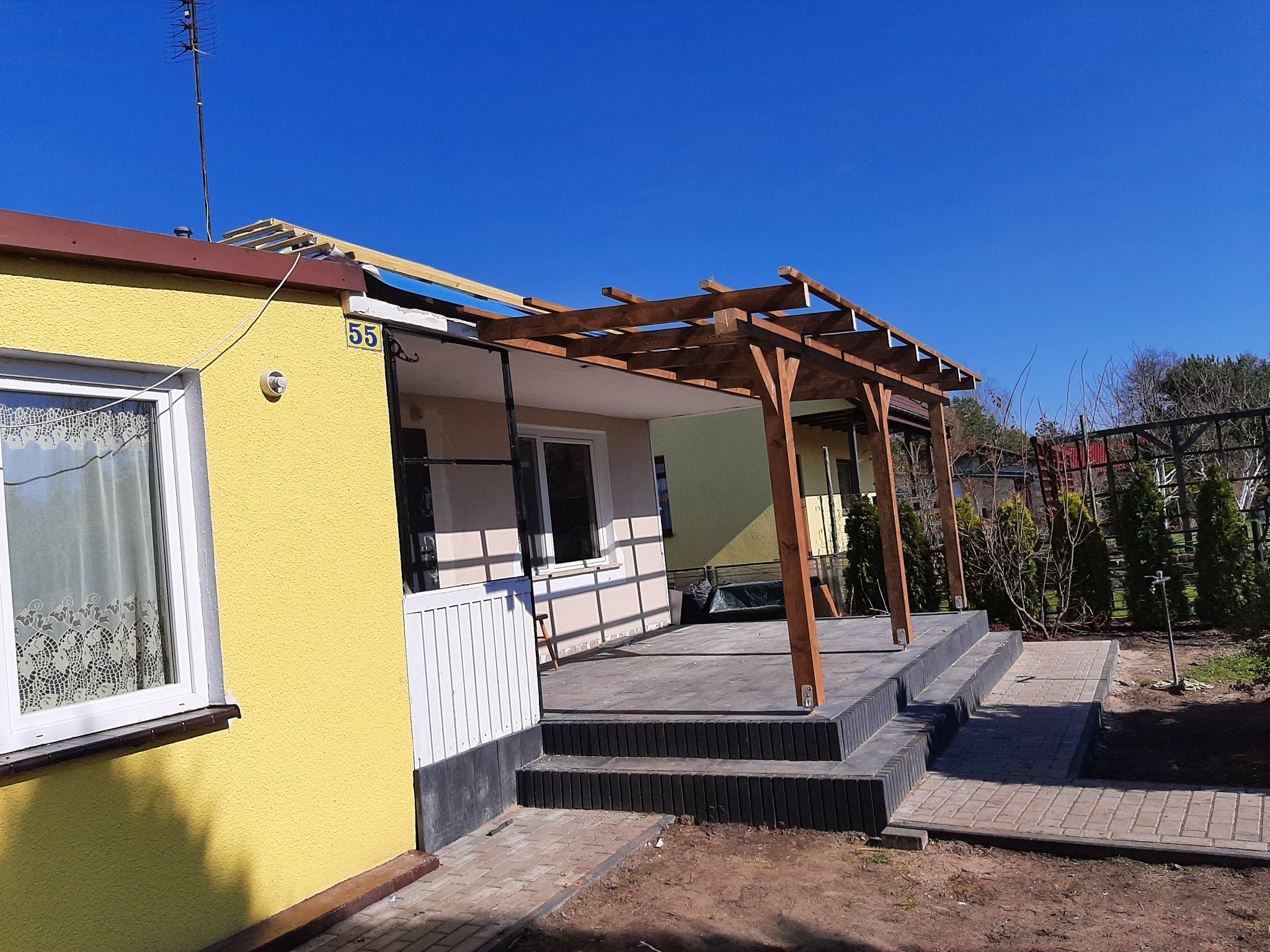 Żółty dom z numerem 55 i drewnianą pergolą w trakcie budowy, widoczne krokwie i częściowo ułożone deski dachowe, obok taras z szarych płyt i schodami.