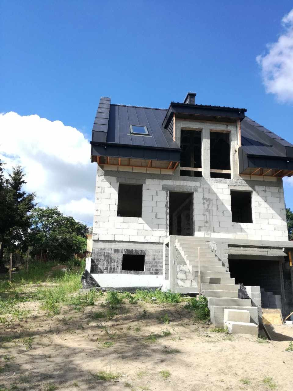 Dwukondygnacyjny dom w stanie surowym otwartym z czarnym dachem, betonowymi schodami wejściowymi i widoczną izolacją fundamentów od strony ogrodu.