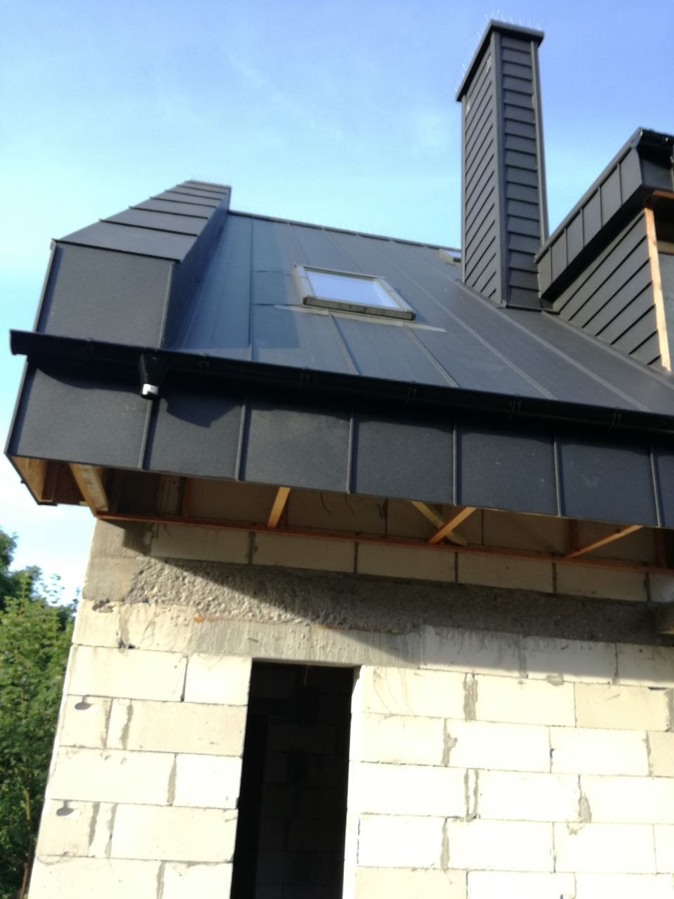 Surowy stan budynku z szarego betonu komórkowego z zamontowanym, grafitowym dachem z blachy na rąbek stojący, z oknem dachowym i kominem, widoczne drewniane elementy konstrukcji dachu.