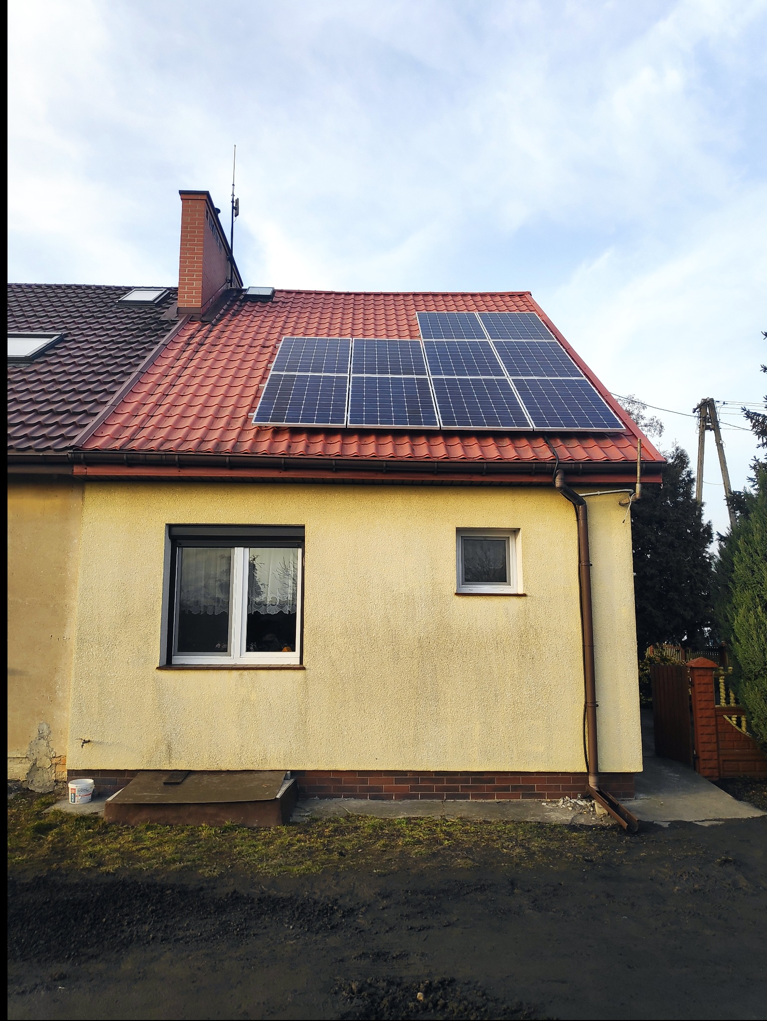 Żółty dom jednorodzinny z czerwoną dachówką, na której zamontowano panele słoneczne; widoczny komin z anteną oraz okna z roletami.