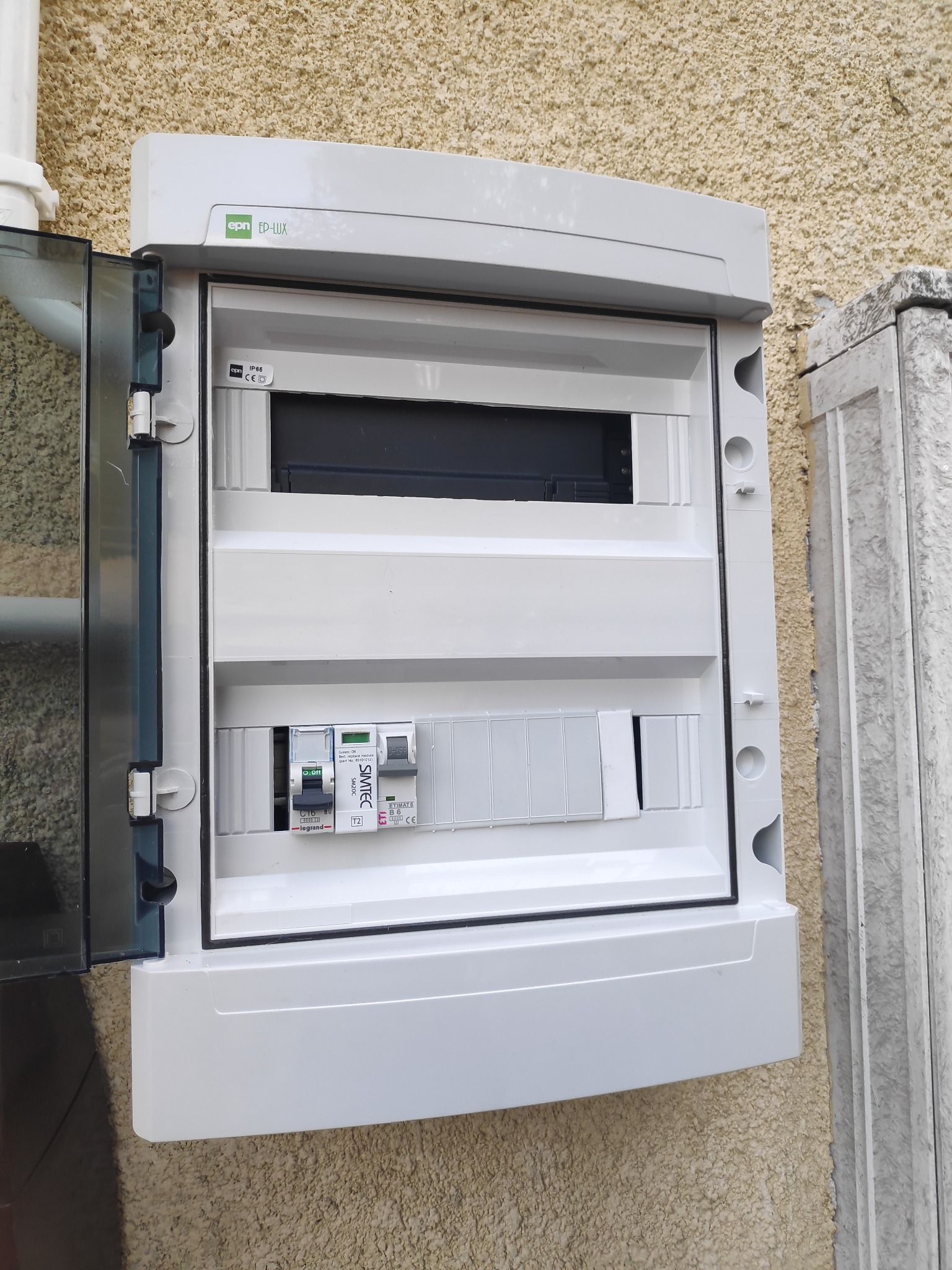 Otwarta skrzynka rozdzielcza z zainstalowanymi bezpiecznikami i miejscem na dodatkowe moduły, zamontowana na zewnętrznej ścianie budynku o teksturowanej powierzchni.