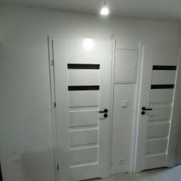 Piorun Piotr Szojda - Dwa białe, nowoczesne drzwi wewnętrzne z czarnymi poziomymi wstawkami i klamkami, zamontowane w jasnym pomieszczeniu w trakcie wykańczania.