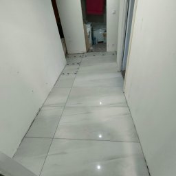 Piorun Piotr Szojda - Jasne, duże płytki podłogowe w korytarzu, częściowo ułożone, z niebieskimi krzyżykami dystansowymi. W tle widoczne wejście do łazienki z pralką.