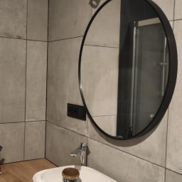 Piorun Piotr Szojda - Nowoczesna łazienka z okrągłym lustrem, umywalką na drewnianym blacie i szarą płytką. Widoczna lampa nad lustrem i fragment pralki. Umywalka zabezpieczona folią.